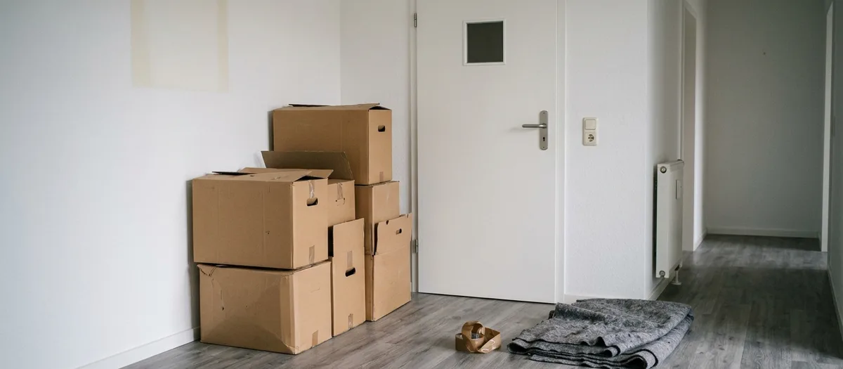 Gestapelte Umzugskartons an der Wand im Flur einer geräumten Wohnung, daneben eine Rolle Klebeband und eine gefaltete Möbeldecke
